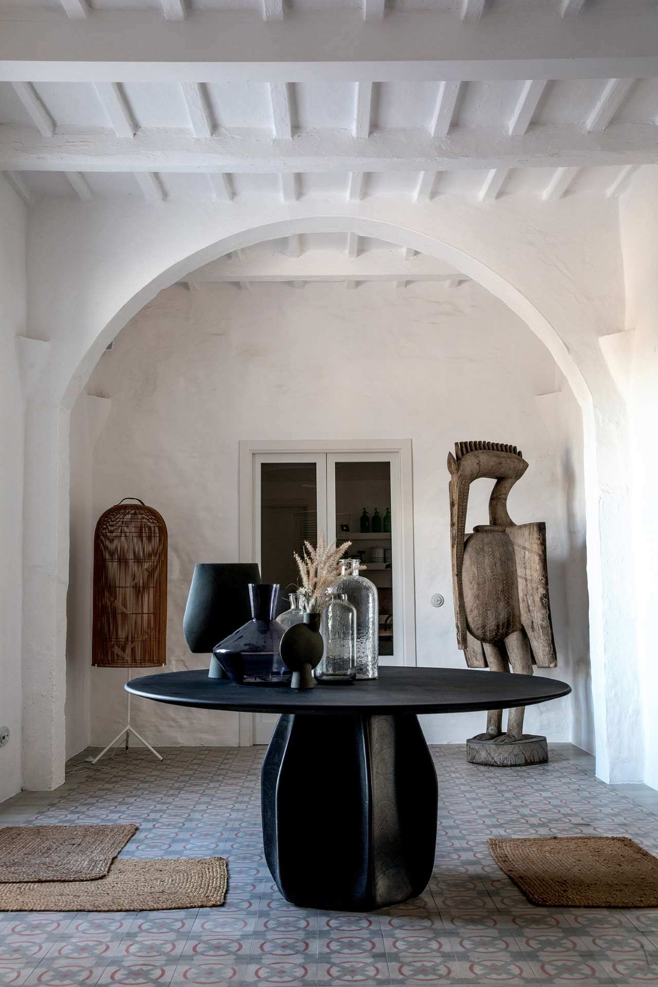 Es Bec d’Aguila — Es Bec d'Aguila, Balearic Islands, Spain — Arched gallery with black pedestal table, sculptural wooden figures, woven textiles, and exposed beam ceiling in whitewashed Mediterranean interior.