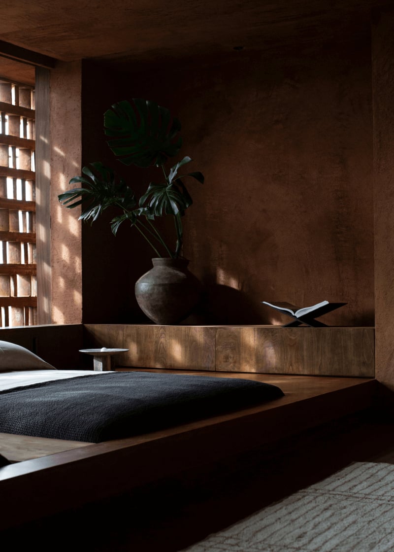 Further Hotel, Asia — Minimalist bedroom with dark wood platform bed, concrete vase with green plant, latticed window casting geometric light shadows on terracotta walls, open book on nightstand
