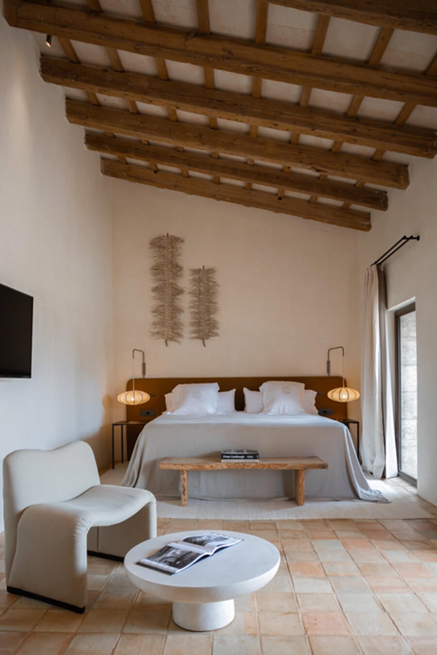 Son Vell, Balearic Islands, Spain — Minimalist bedroom with exposed wooden ceiling beams, linen upholstery chair, sculptural nightstands, and terracotta tile flooring.