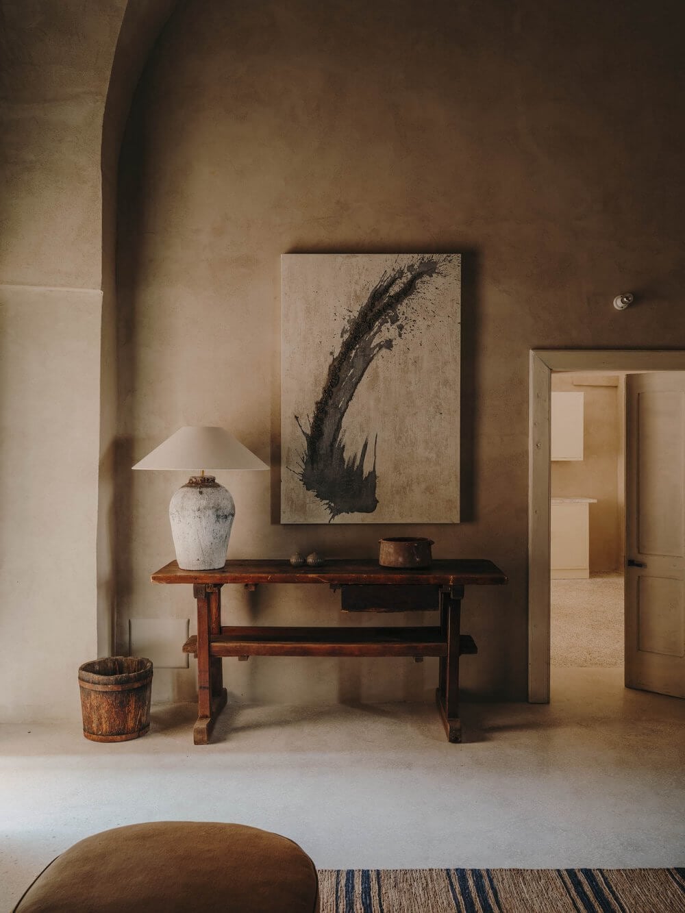 Casa Soleto, Europe — Minimalist entryway with terracotta table, textured plaster walls, abstract charcoal artwork, ceramic lamp, and wooden vessel in warm natural light.