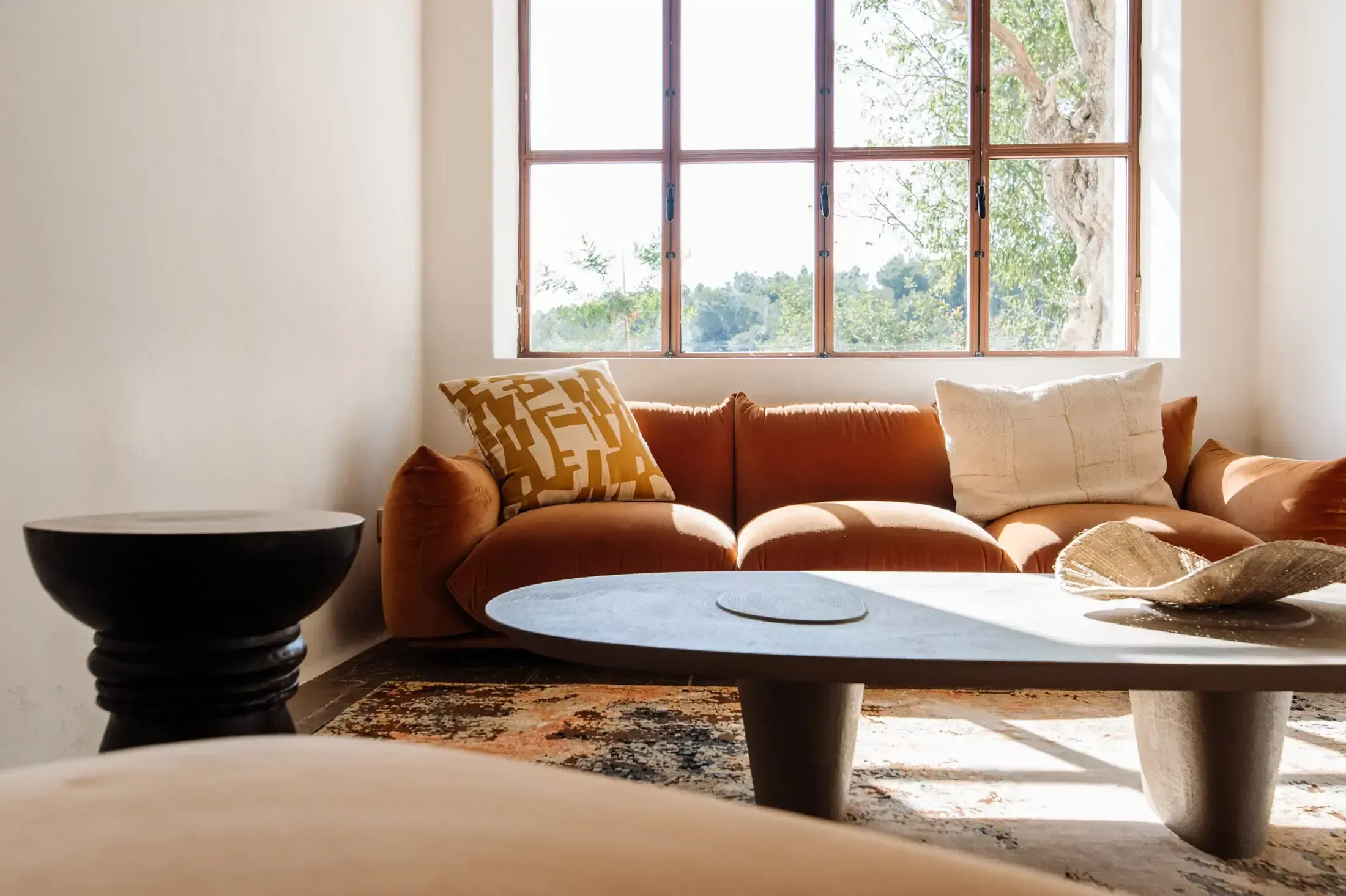 Finca La Inika, Balearic Islands, Spain — Terracotta sofa with geometric pillows positioned before large windows overlooking green landscape, concrete oval table, natural light, contemporary luxury interior.