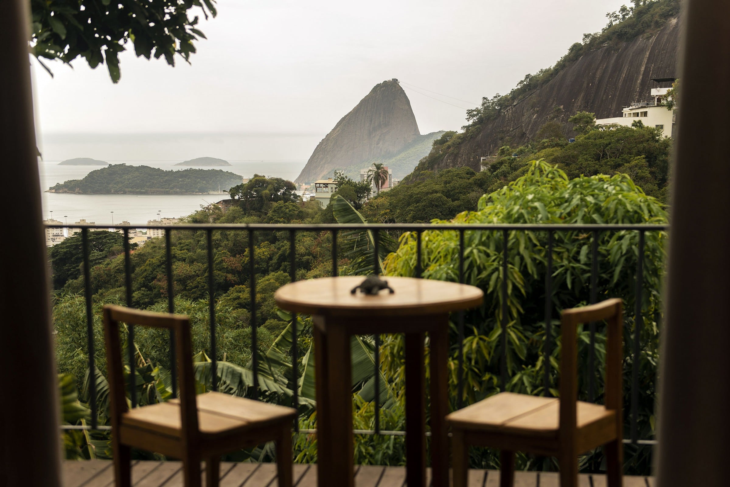 Chez Georges, South America — Coastal terrace with wooden chairs and table overlooking Sugar Loaf Mountain, lush tropical vegetation, and calm bay waters under overcast sky.