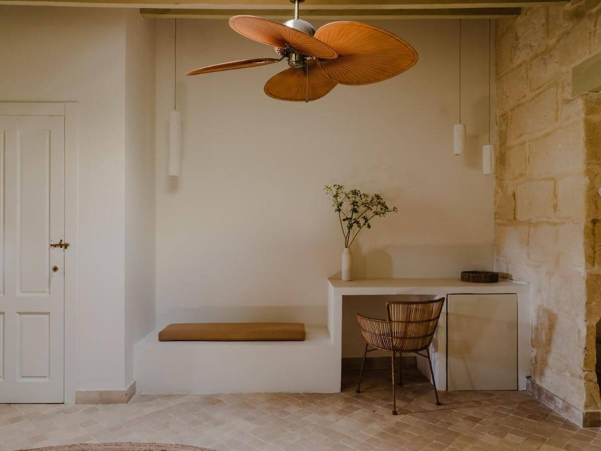 Amagatay, Balearic Islands — Minimalist bedroom with wooden ceiling fan, white built-in bench, woven chair, terracotta tile floor, and preserved stone wall detail.