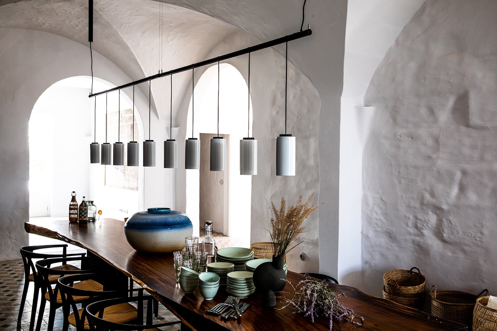Es Bec d’Aguila — Es Bec d'Aguila, Balearic Islands, Spain — Dining room with arched windows, dark wood table, cylindrical pendant lights, ceramic vessels, and whitewashed stone walls.