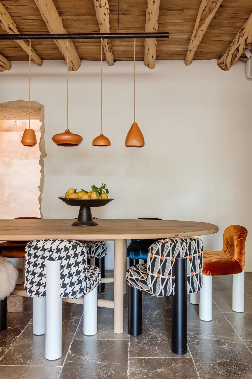Finca La Inika, Balearic Islands, Spain — Rustic wooden beams frame copper pendant lights above oval dining table with patterned stools and fruit bowl.