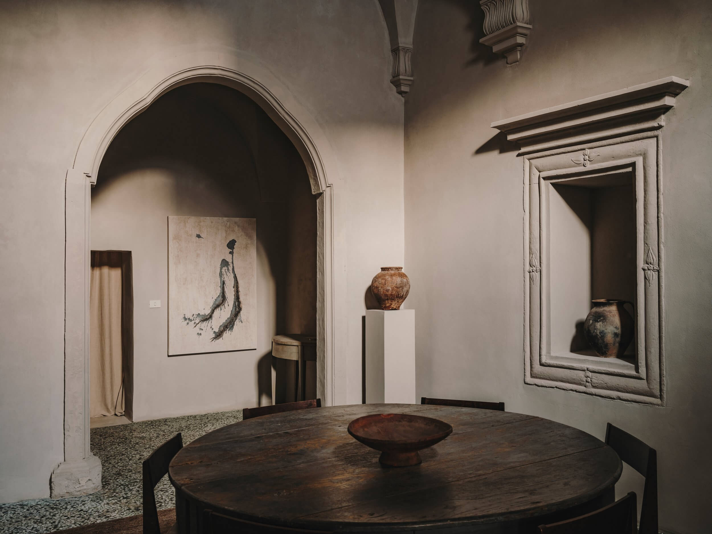 Casa Soleto, Europe — Arched gallery with round wooden table, terracotta vessels on pedestals, minimalist line drawing, travertine floors, classical architecture.