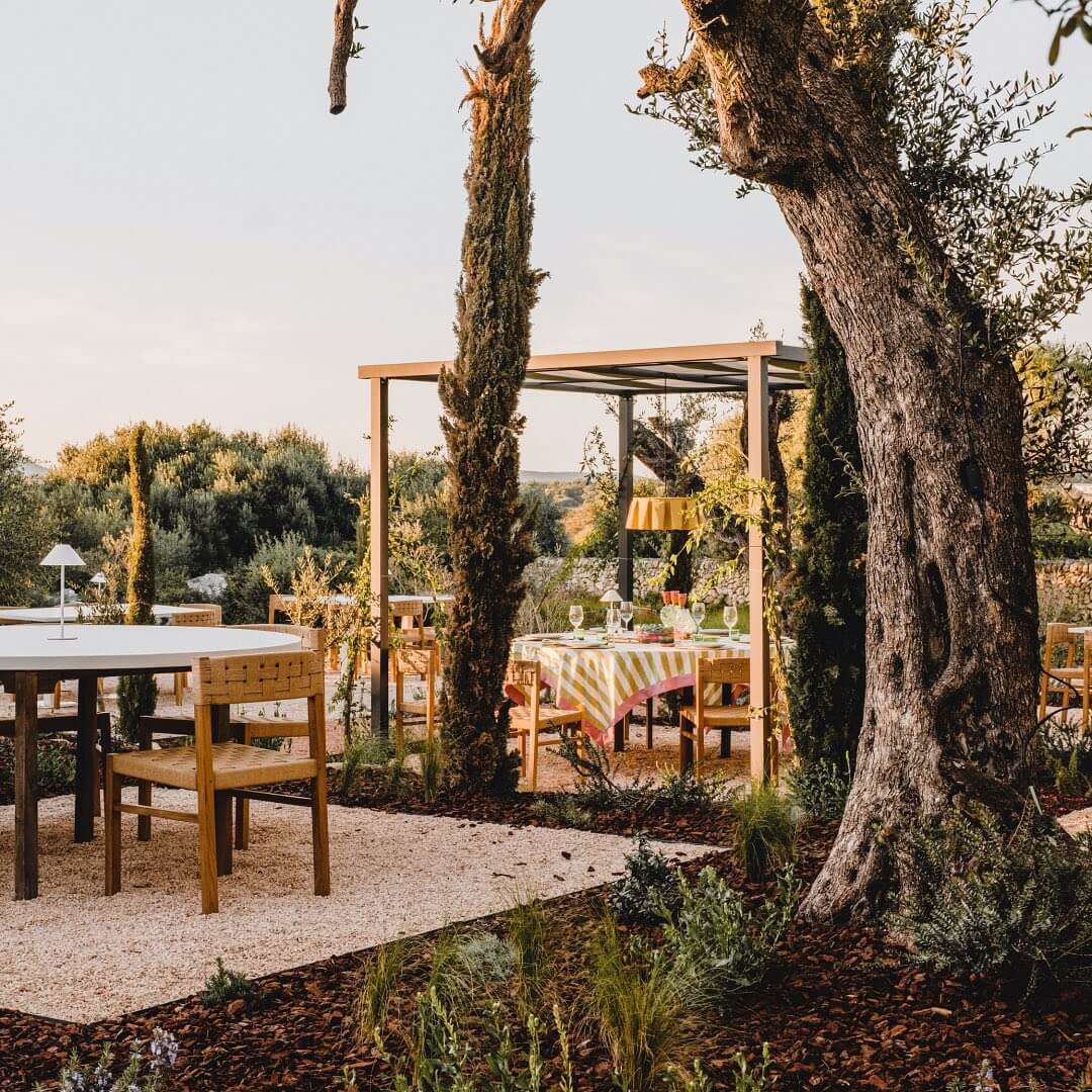 Amagatay, Balearic Islands — Wooden pergola and dining tables beneath ancient olive trees on gravel terrace at sunset, Mediterranean landscape hospitality design.