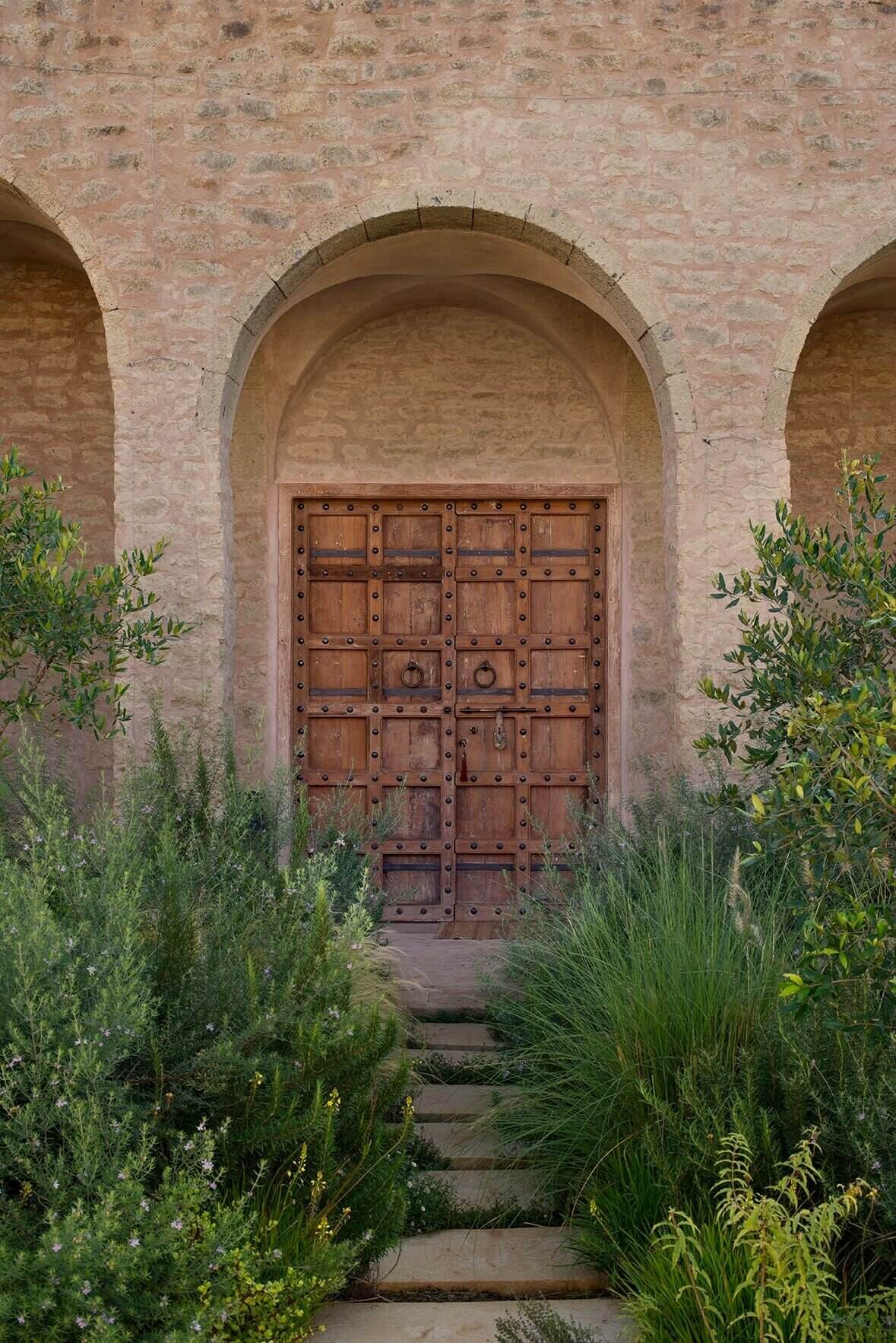 Arijiju Retreat, Africa — Arched stone doorway with studded wooden door flanked by Mediterranean plants, warm earth tones, minimalist luxury architecture.