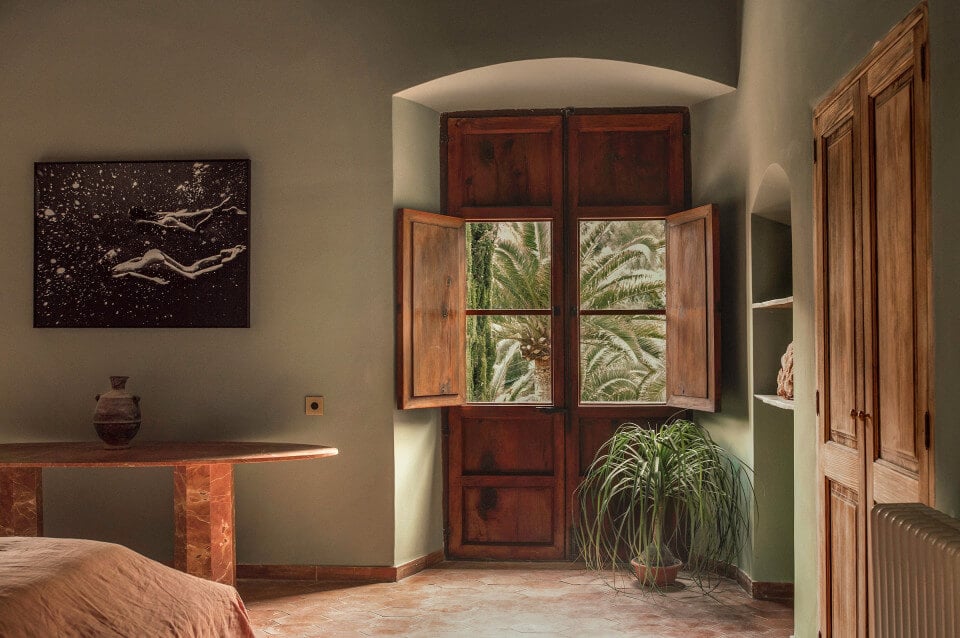 Hotel Corazón, Balearic Islands, Spain — Sunlit bedroom with terracotta shutters, arched niche, minimalist furnishings, palm fronds beyond window, earthy Mediterranean aesthetic.