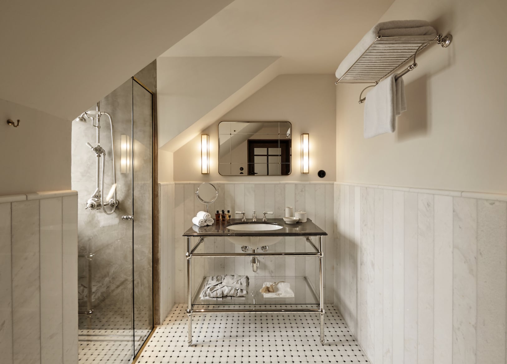 Hotel Sanders, Europe — Attic bathroom with marble-clad walk-in shower, polished nickel console sink, vintage-style fixtures, and geometric floor tiles beneath angled ceiling.