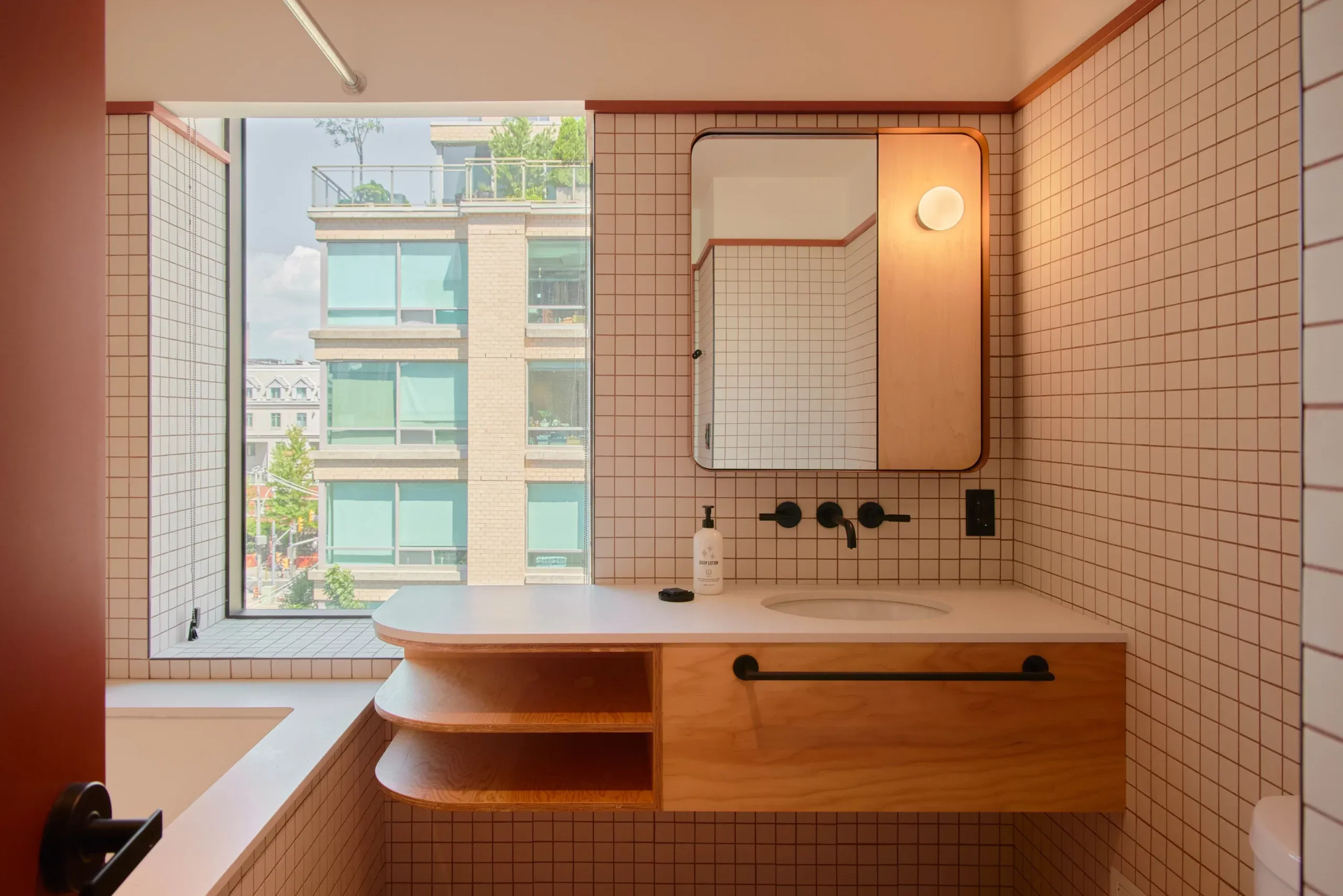 Ace Hotel Toronto, Canada — Modern bathroom with terracotta tile, warm wood vanity, rounded mirror, black fixtures, and floor-to-ceiling windows overlooking city streetscape.