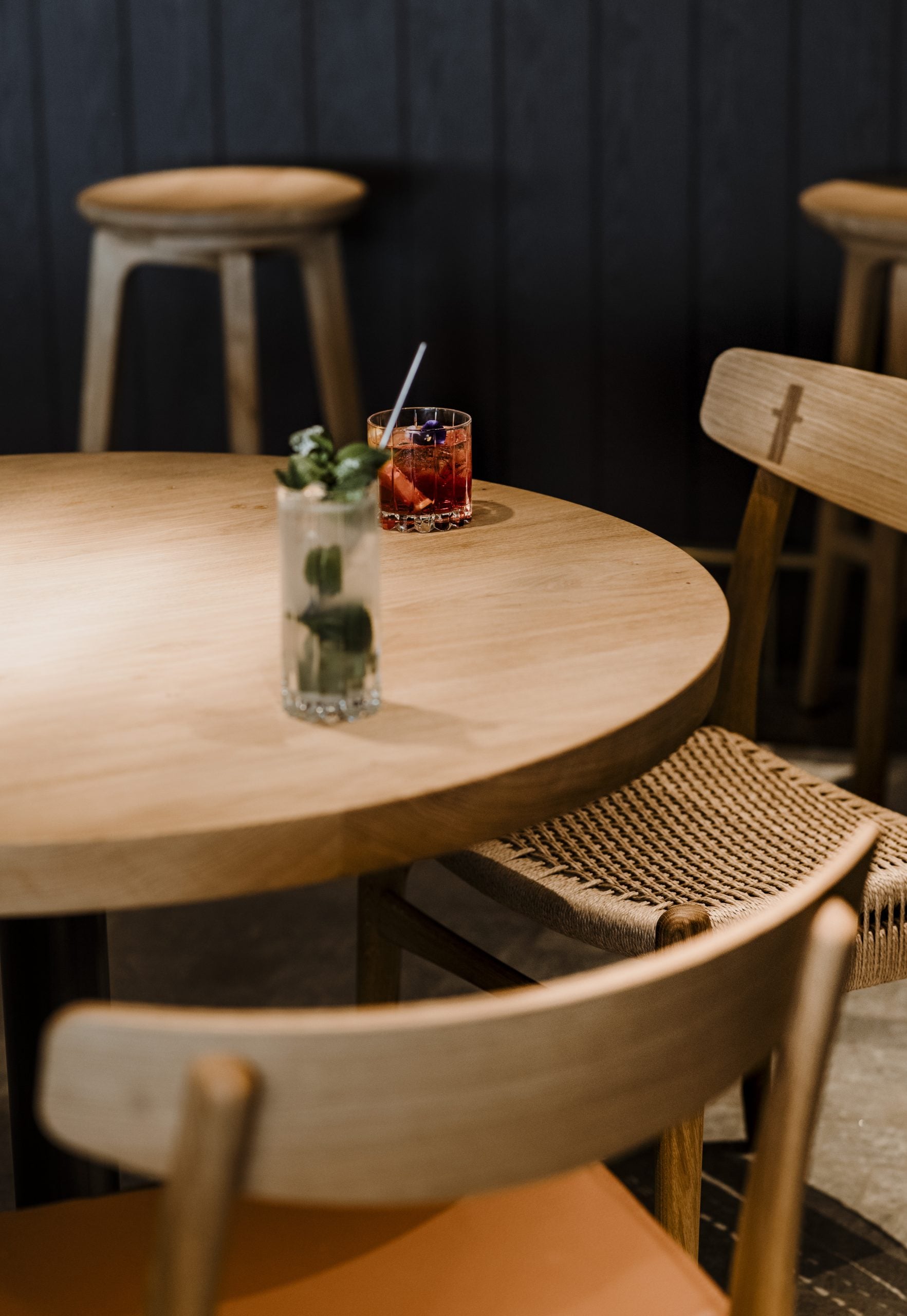 Casa di Langa, Europe — Light wood round table with woven chairs, reddish cocktail with straw, fresh herbs in glass, dark curtained backdrop, minimalist hospitality interior design.