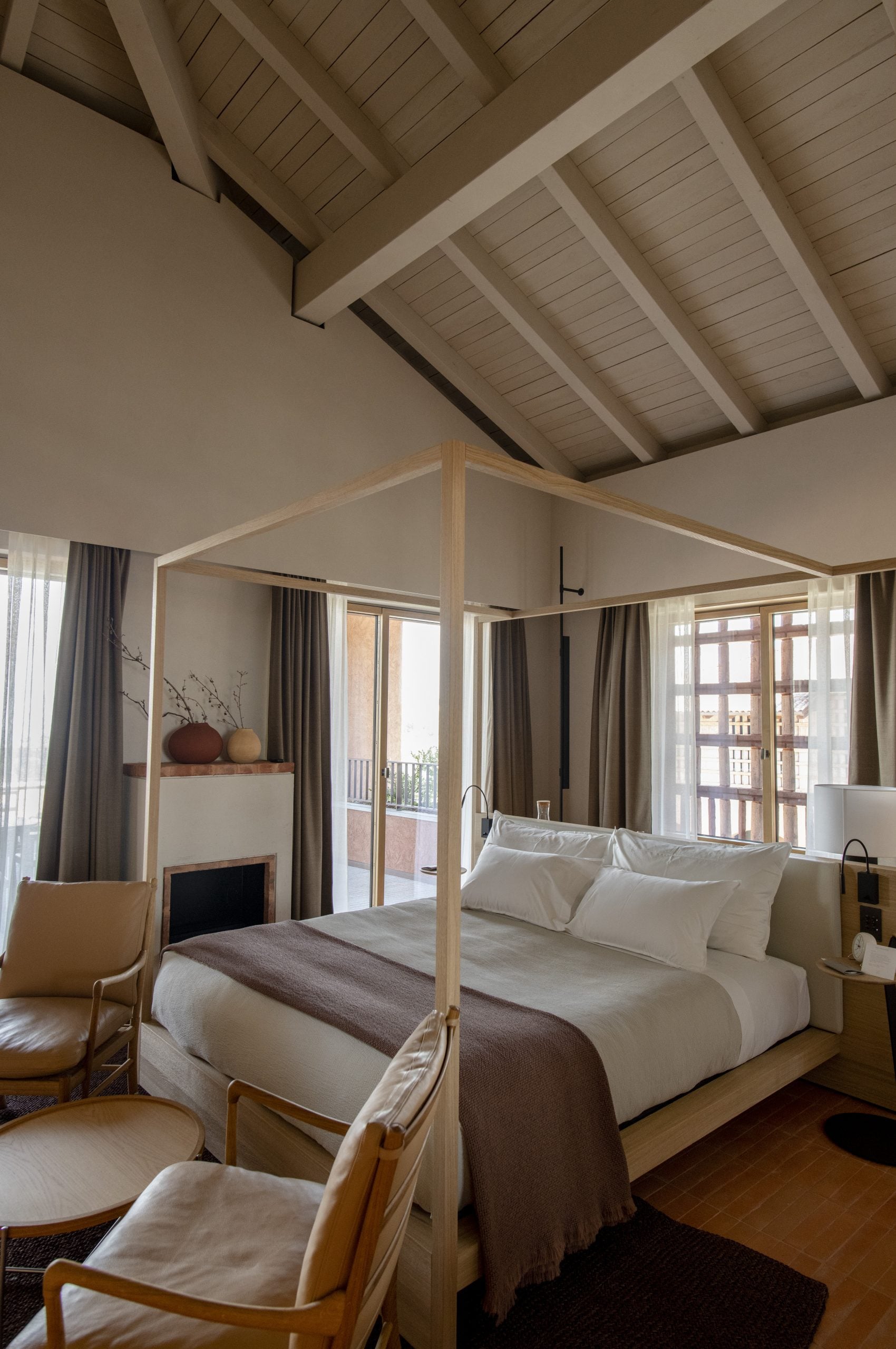 Casa di Langa, Europe — Minimalist bedroom with natural wood canopy bed, exposed beam ceiling, floor-to-ceiling windows, neutral linens, and modern fireplace.