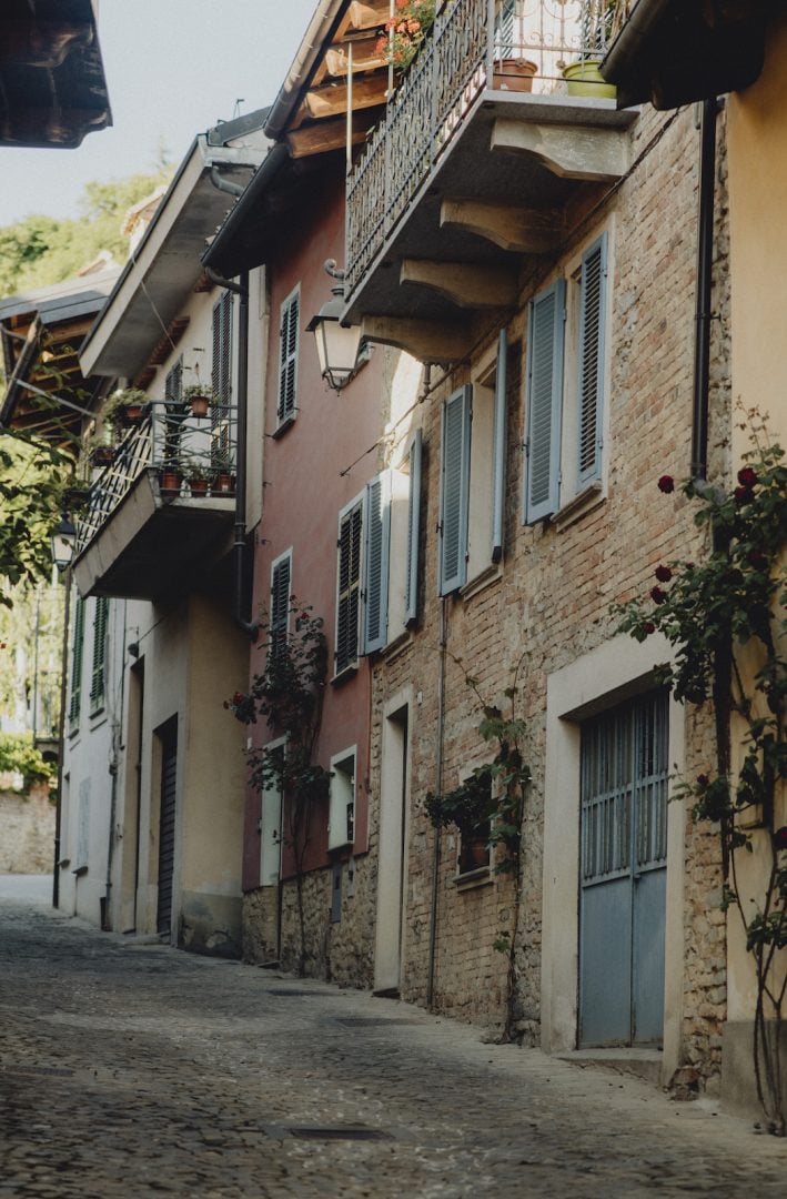 Casa di Langa, Europe — Narrow cobblestone alley lined with terracotta and stone buildings, green shutters, ivy-covered walls, wooden balconies casting shadows.