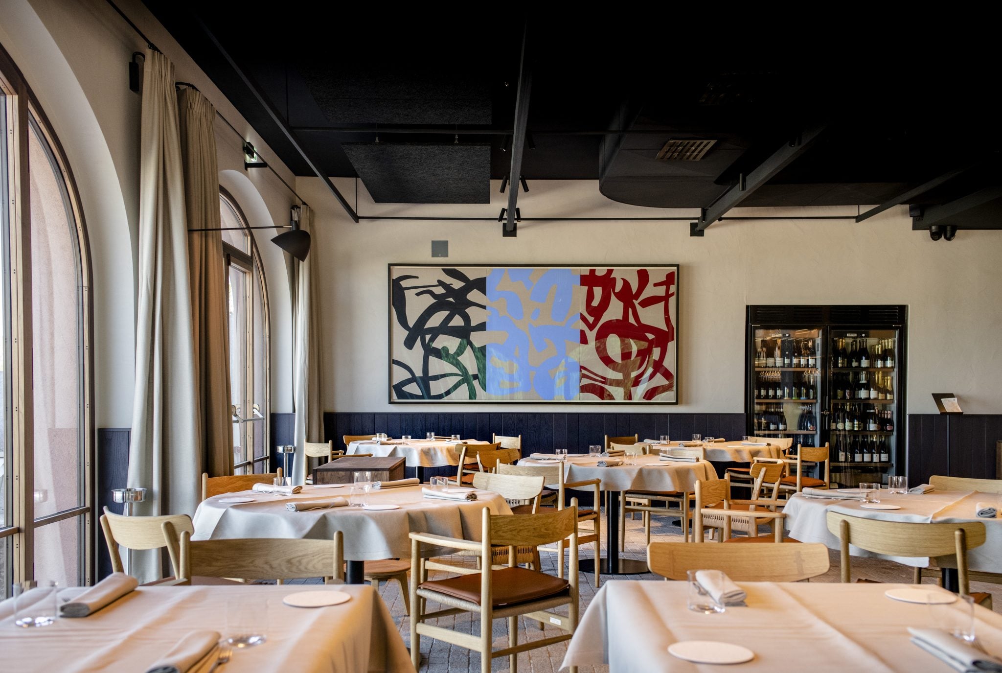 Fàula Ristorante, Europe — Contemporary dining room with natural wood chairs, white tablecloths, large colorful abstract triptych, dark wainscoting, floor-to-ceiling windows, wine displays, and minimalist ceiling details.