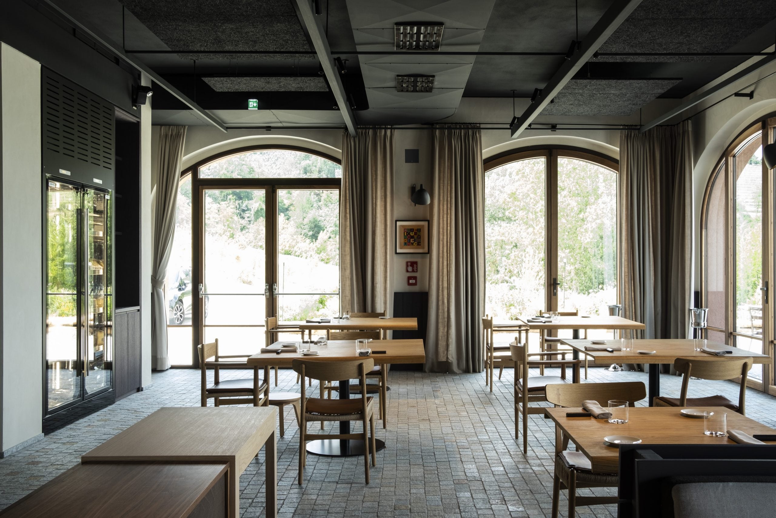 Casa di Langa, Europe — Modern dining room with arched windows, wooden tables, natural light, and views of forested hillside.