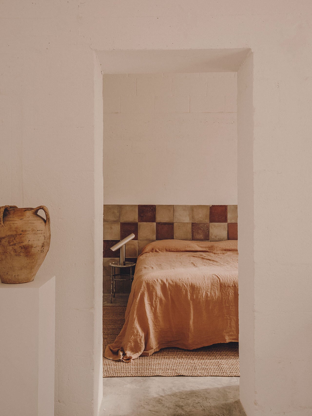 Borgo Gallana, Europe — Minimalist bedroom with terracotta bedding, checkered tile backsplash, neutral plaster walls, and earthenware vessel detail.