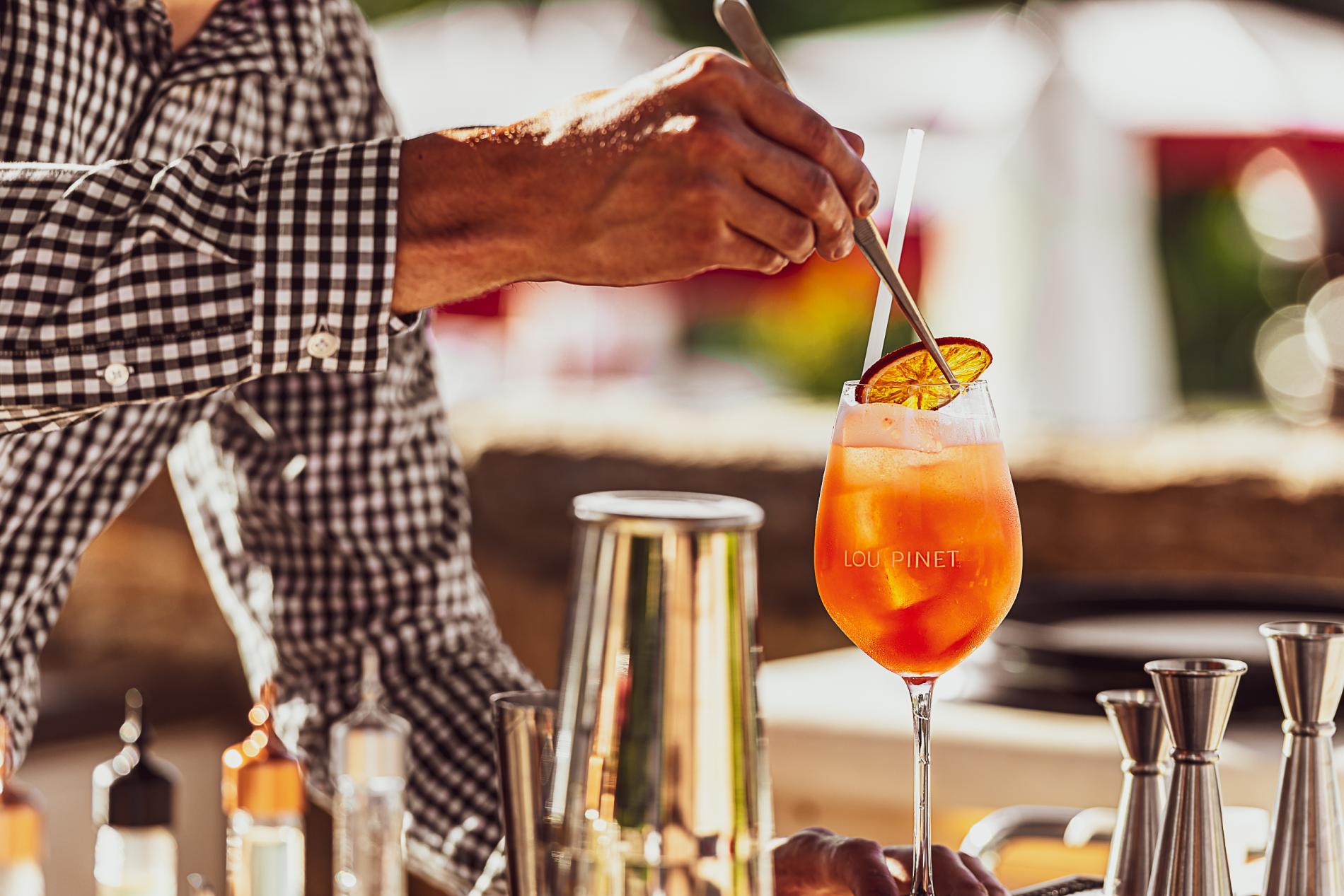 Hôtel Lou Pinet, Europe — Bartender stirring aperitif with orange slice in stemware, natural light, luxury hospitality service.