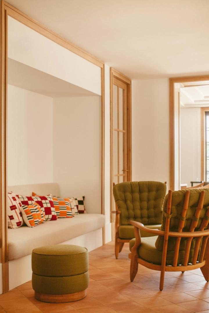 Hôtel Lou Pinet, Europe — Minimalist lounge with cream built-in sofa, olive upholstered chairs, terracotta tile flooring, and natural wood framing in warm Mediterranean light.