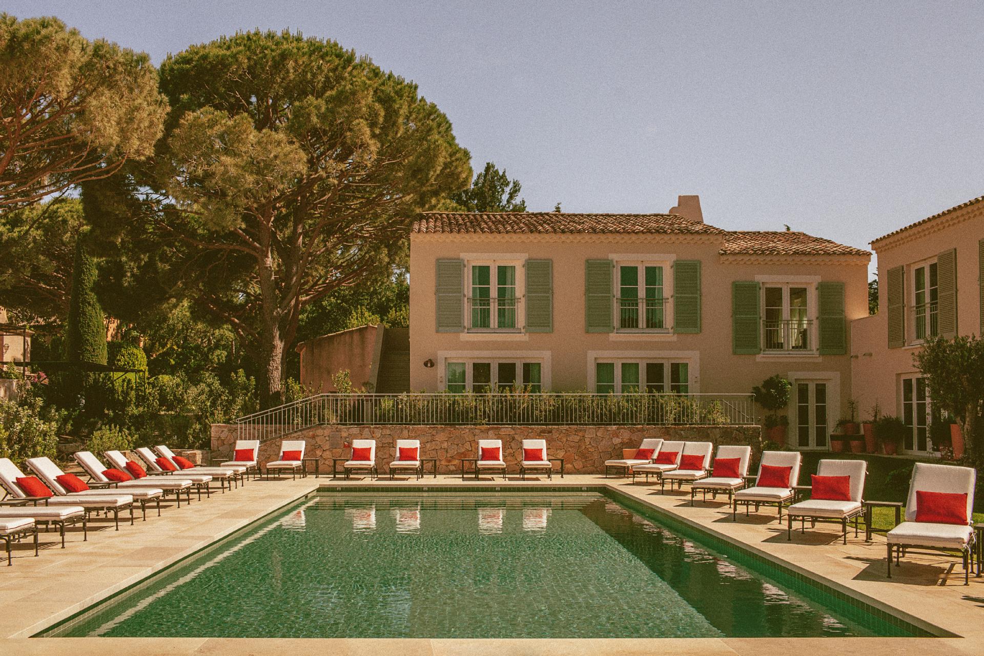 Hôtel Lou Pinet, Europe — Poolside terrace with stone building, green shutters, loungers with red cushions, and Mediterranean pine trees in afternoon light.