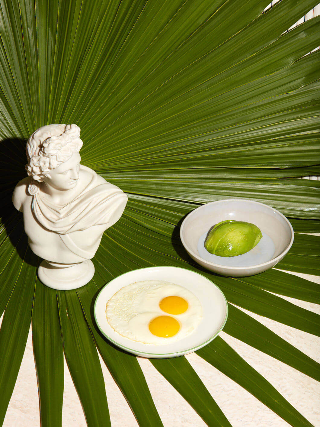 Palm Heights, North America — Minimalist still life with classical bust, fried eggs, lime, and palm fronds on white surface, bright natural light.