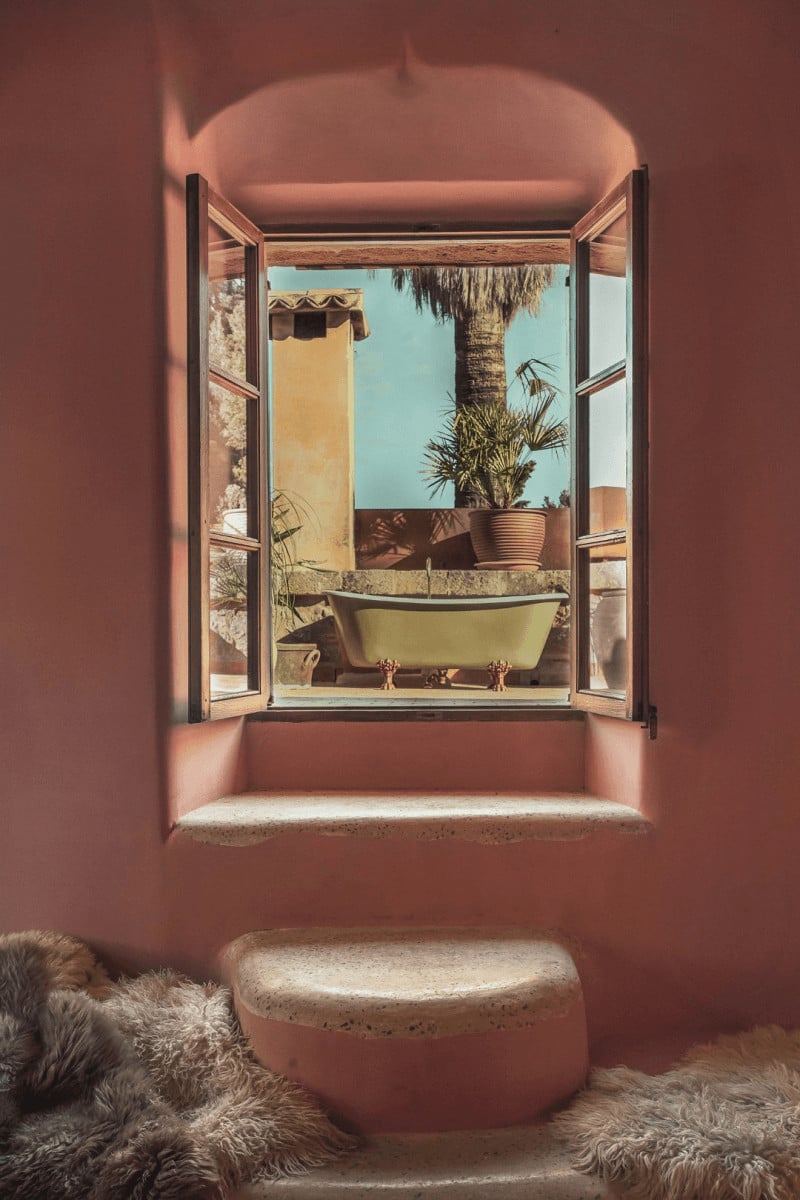 Hotel Corazón, Balearic Islands — Terracotta interior with open wooden shutters framing yellow bathtub, palm tree, and clear blue sky through window.