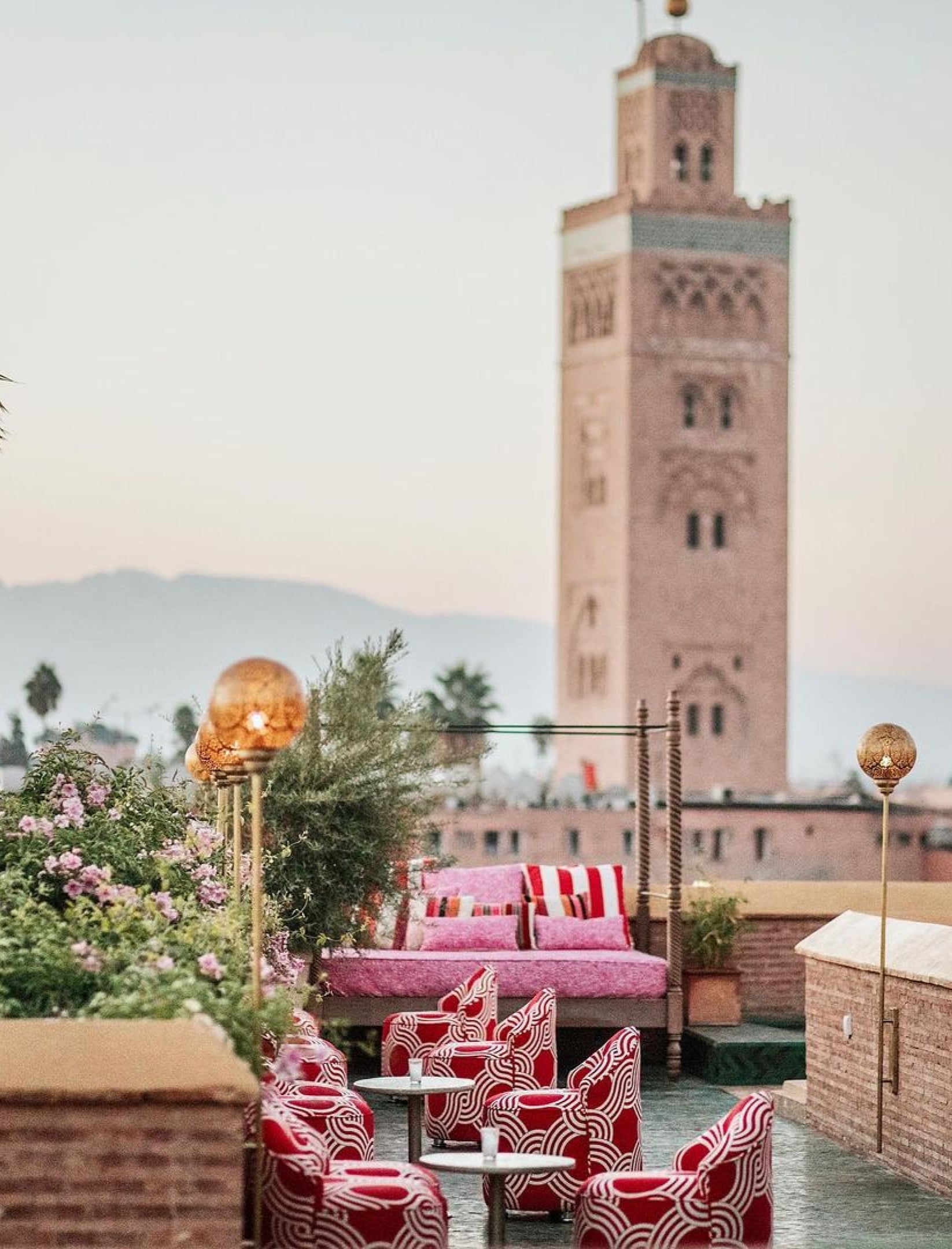 El Fenn Rooftop Bar, Marrakech, Morocco — Pink seating with geometric red cushions overlooks Koutoubia Mosque minaret at sunset, mountains beyond.