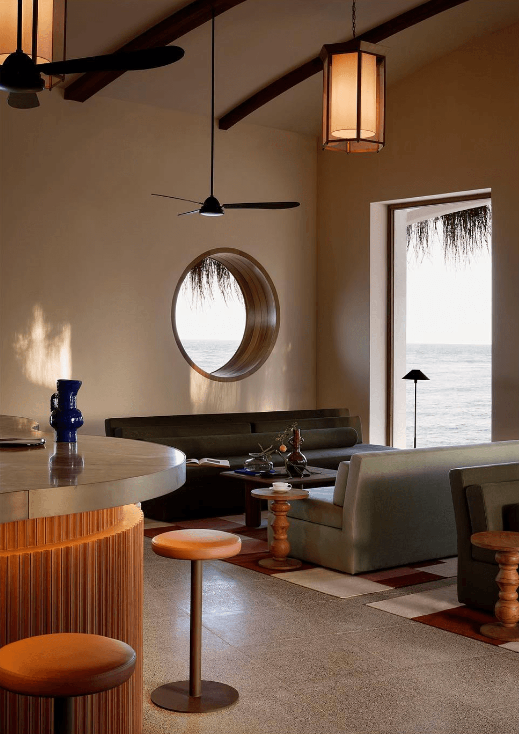 Kisawa Sanctuary, Africa — Coastal lounge with circular porthole window, dark wooden beams, pendant lighting, sage upholstered seating, and ocean views through glass doors.