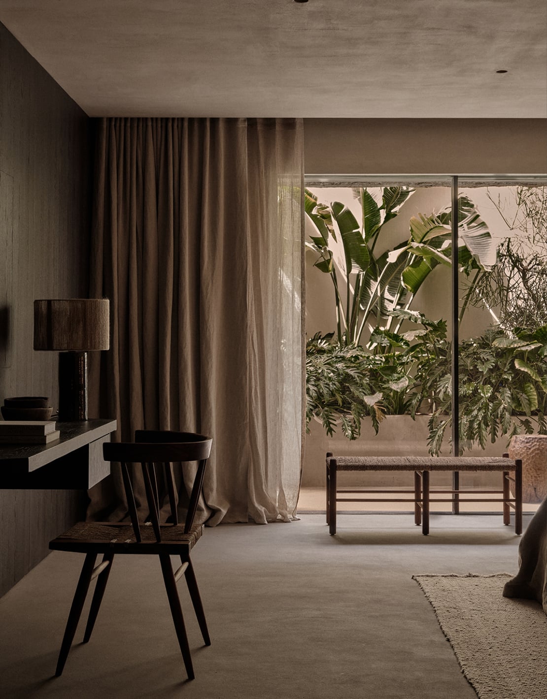En Kyanó, Europe — Minimalist bedroom with concrete ceiling, linen curtains, black wooden furniture, and floor-to-ceiling windows opening onto lush plant-filled terrace.
