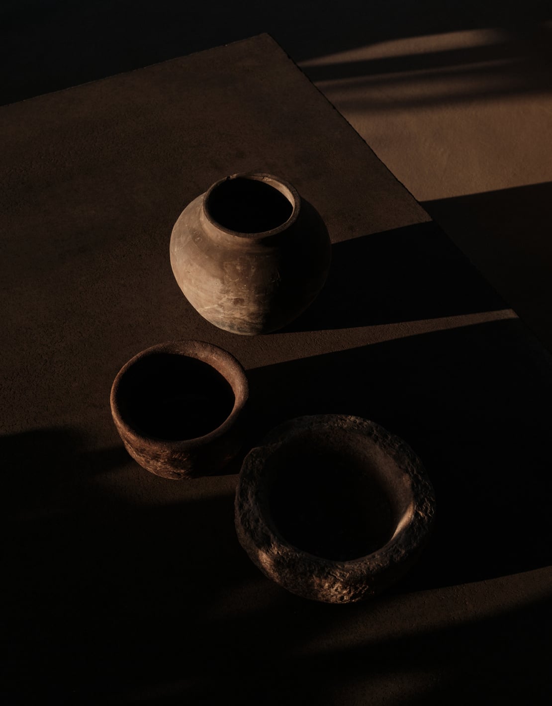 En Kyanó, Europe — Three terracotta vessels arranged on stone floor with dramatic shadow play, warm natural light illuminating earthenware textures and minimalist interior architecture.