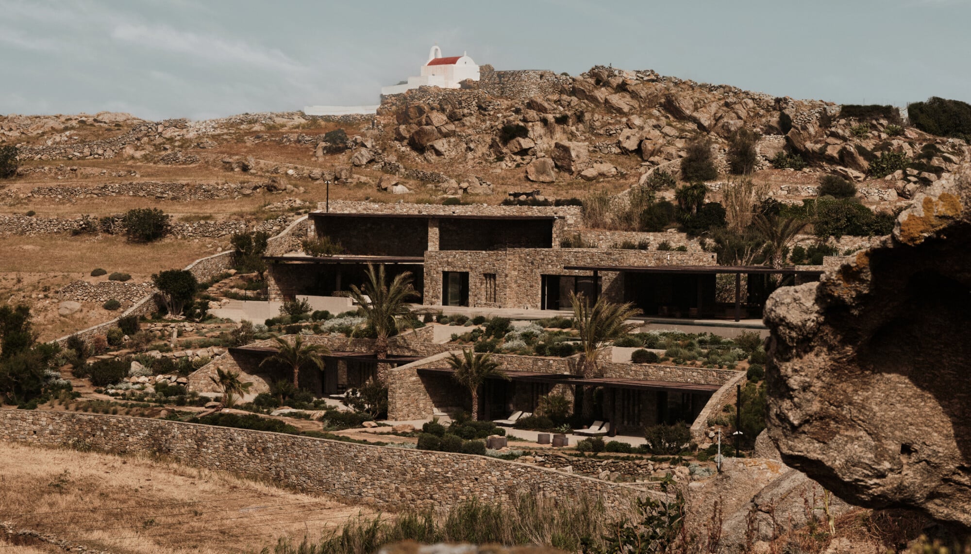 En Kyanó, Europe — Modern stone villa nestled into rocky hillside, featuring minimalist architecture with panoramic windows, desert landscape, and Mediterranean vegetation.