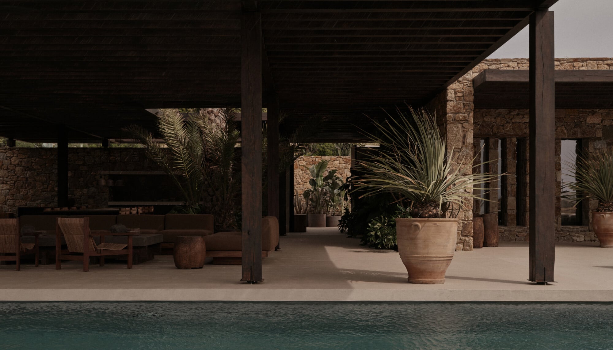 En Kyanó, Europe — Poolside terracotta courtyard with dark wood pergola, stone walls, potted palms, and minimalist lounge seating under Mediterranean sun.