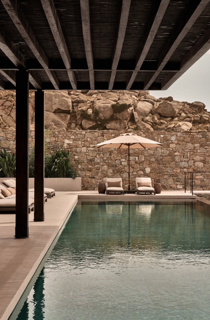 En Kyanó, Europe — Minimalist poolside terrace with stone walls, wooden pergola, loungers, and umbrella beneath dramatic granite boulders.