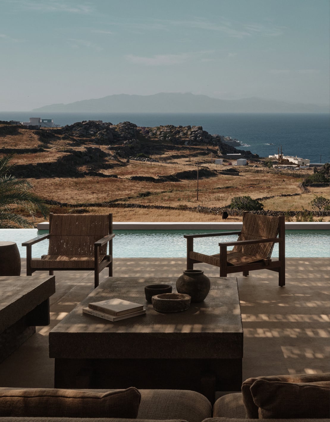 En Kyanó, Europe — Coastal villa terrace with woven lounge chairs, infinity pool, and Mediterranean mountain views across turquoise sea at golden hour.