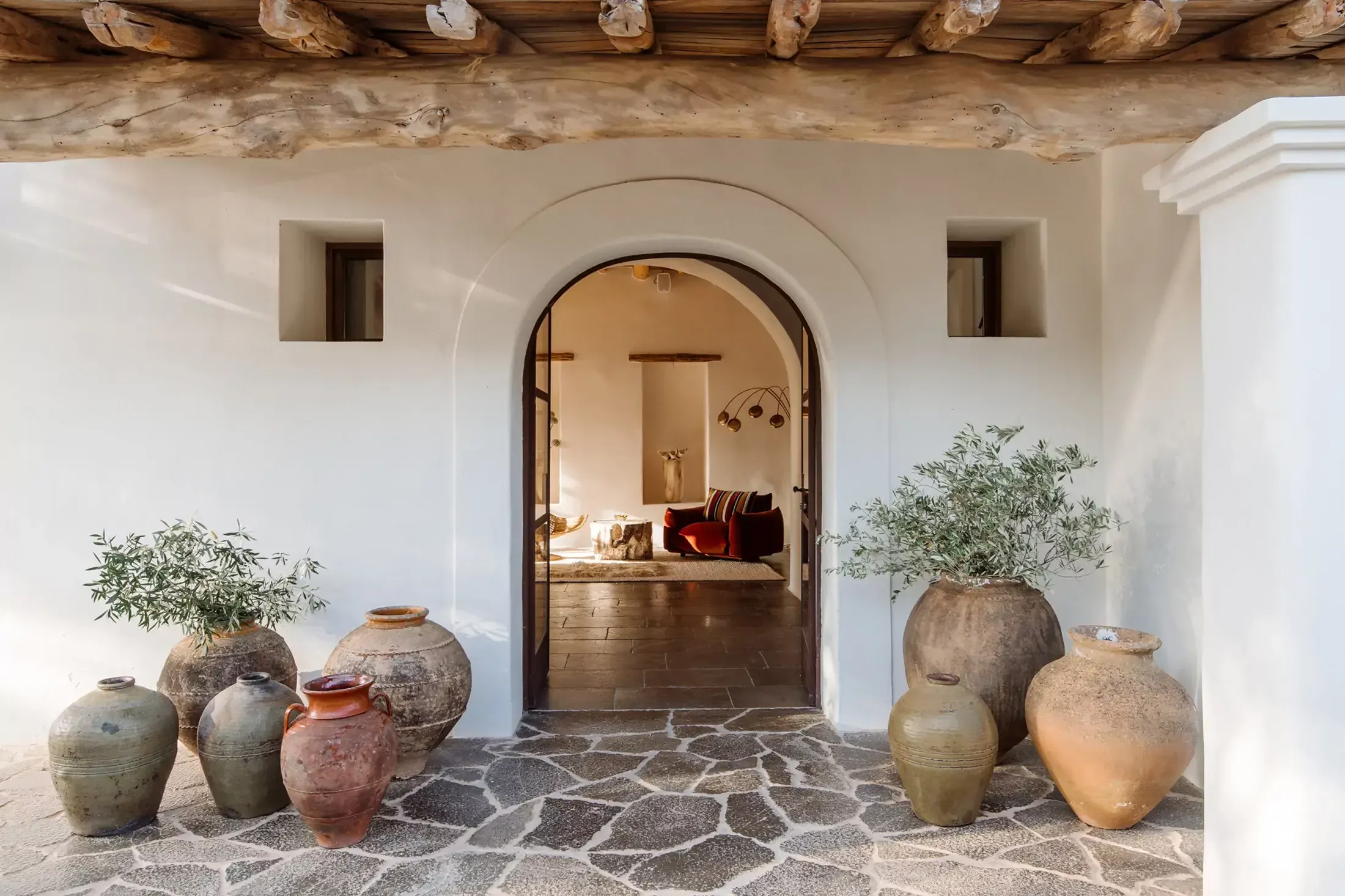 Finca La Inika, Balearic Islands, Spain — Arched doorway with terracotta vessels and olive plants opening to minimalist interior with exposed wooden beams and warm stone flooring.