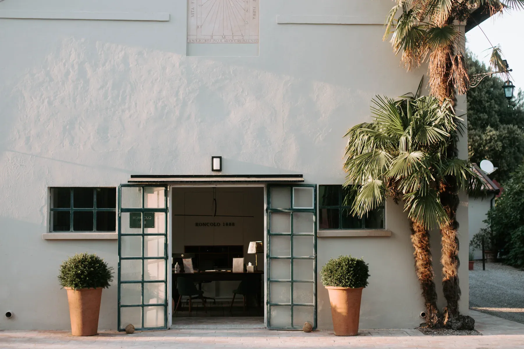 Roncolo 1888, Europe — Minimalist whitewashed façade with teal-framed glass doors, potted boxwoods, and tall palm casting shadows in warm afternoon light.
