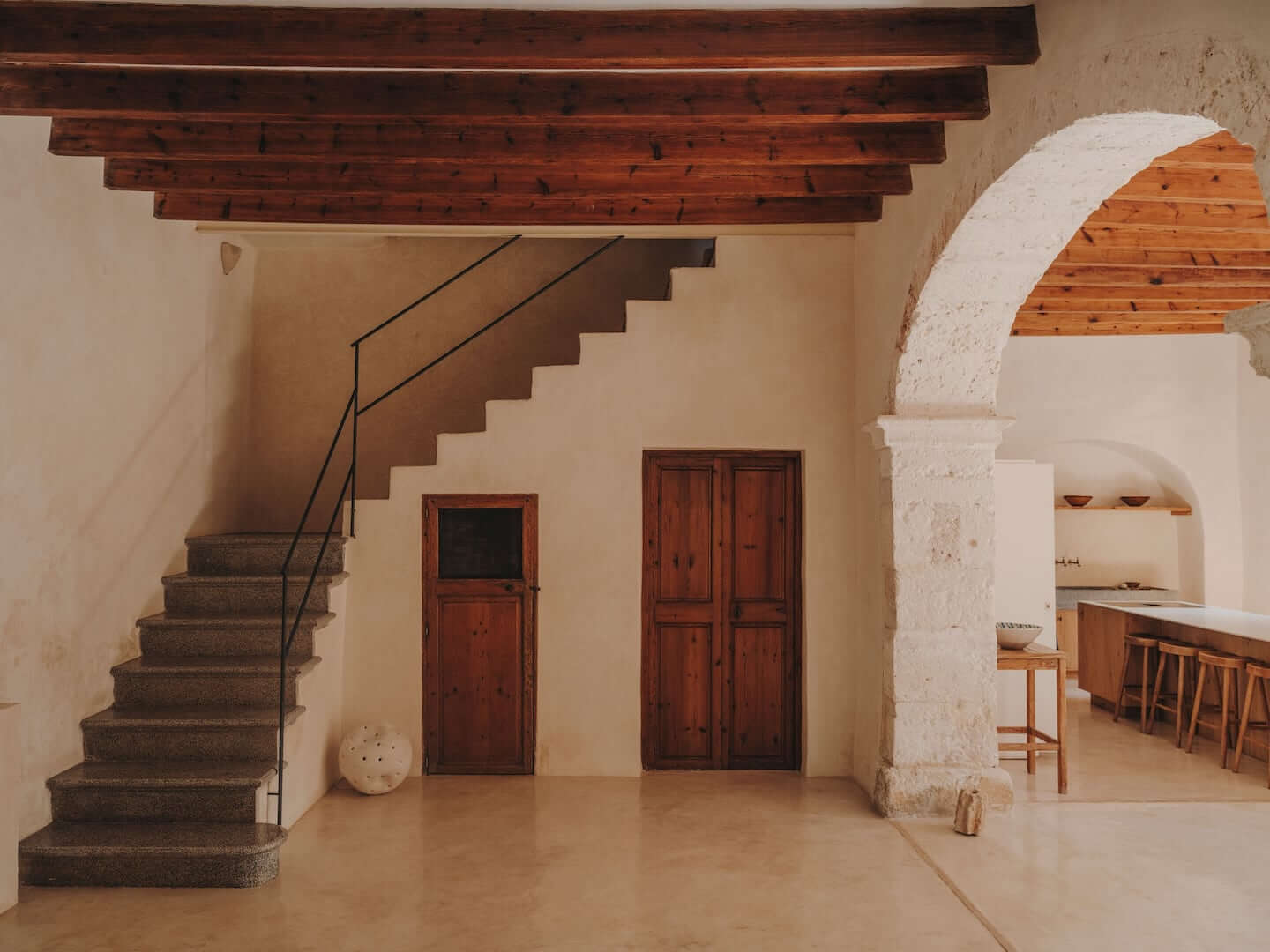 Can Lluïssó, Balearic Islands, Spain — Arched stone doorway, exposed wooden beams, concrete staircase with iron railing in minimalist Mediterranean interior.