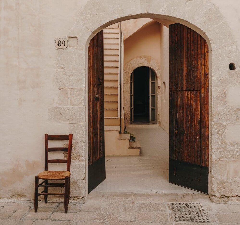 Casa Soleto, Europe — Arched wooden doors opening to stone interior with rustic chair, warm Mediterranean light, minimalist luxury aesthetic.