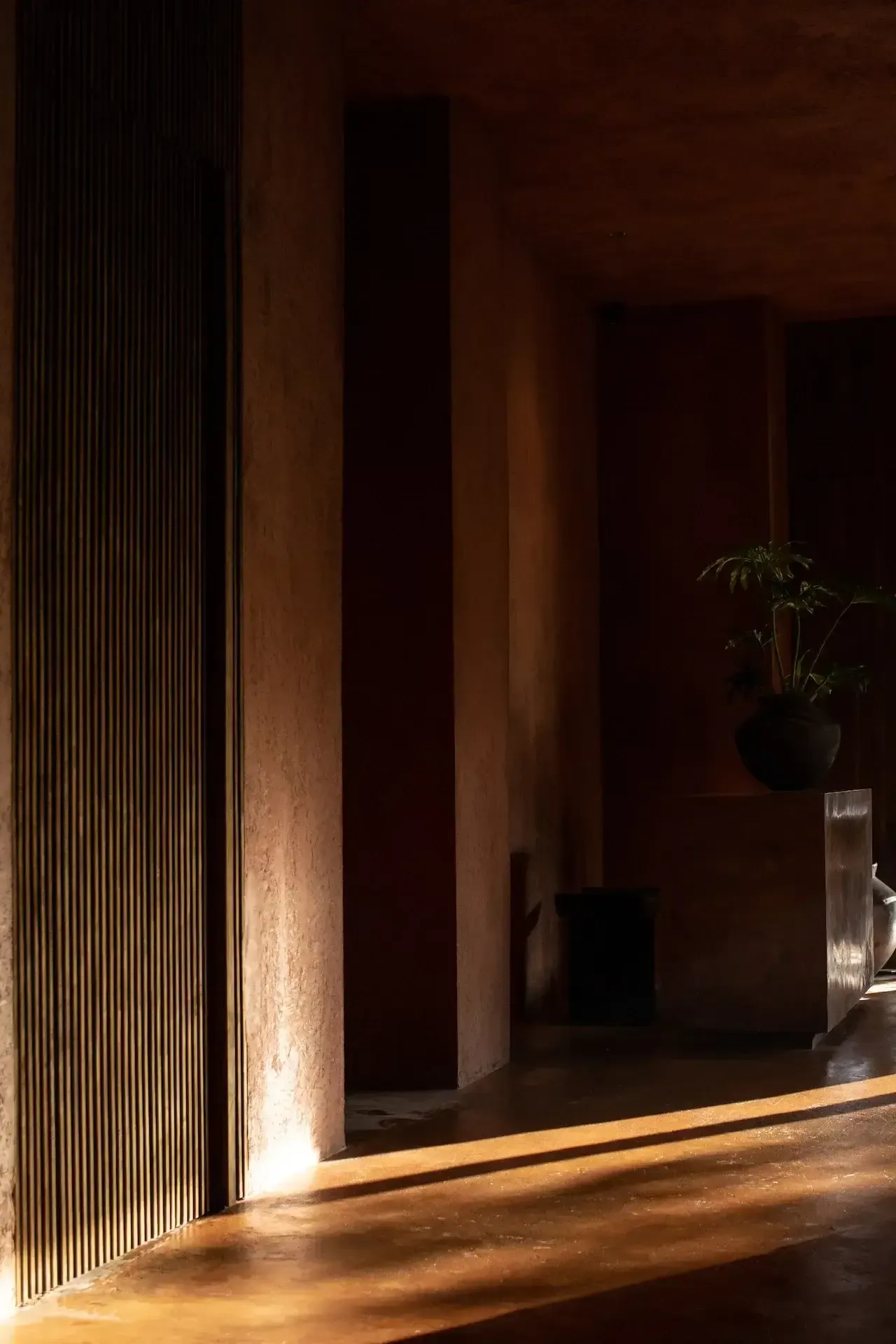 Further Hotel, Asia — Minimalist corridor with ribbed metal wall, warm uplighting, concrete pillars, potted plant, polished floor casting dramatic shadows.