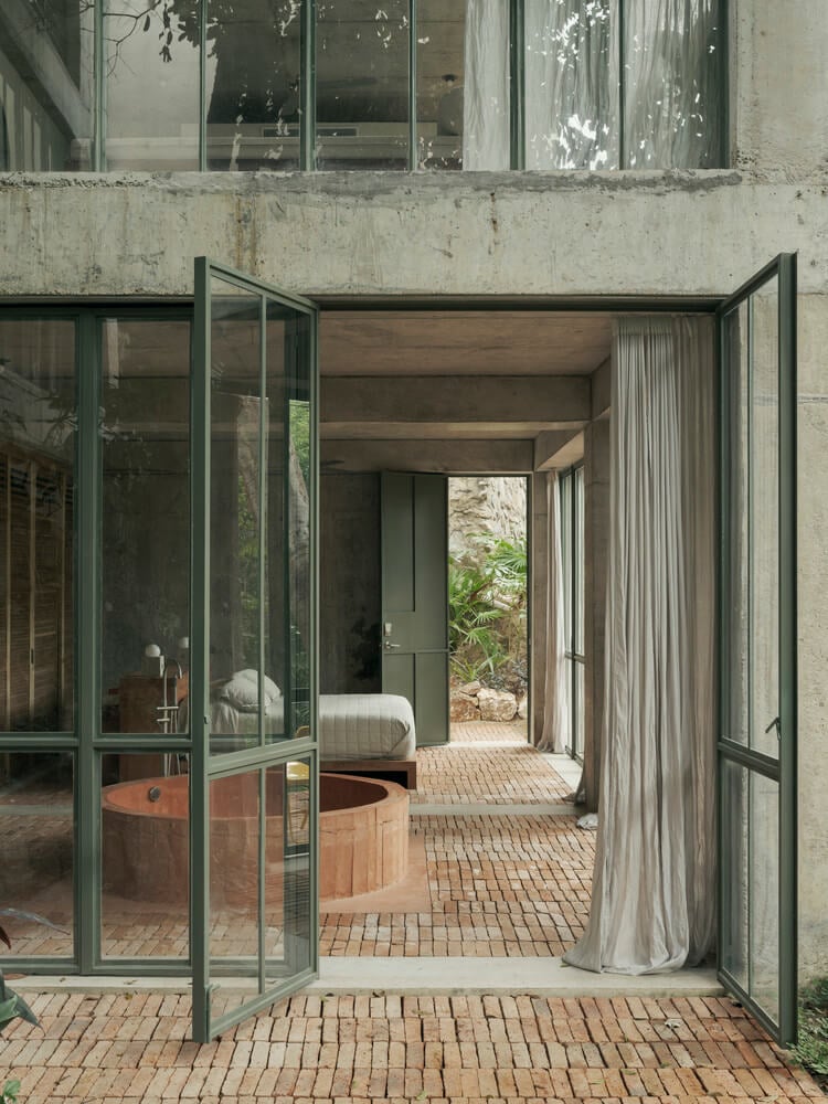 A breezeway at NICO reveals layered concrete architecture, sage-green steel-framed doors, terracotta brick flooring, and a wooden soaking tub glimpsed through glass partitions.