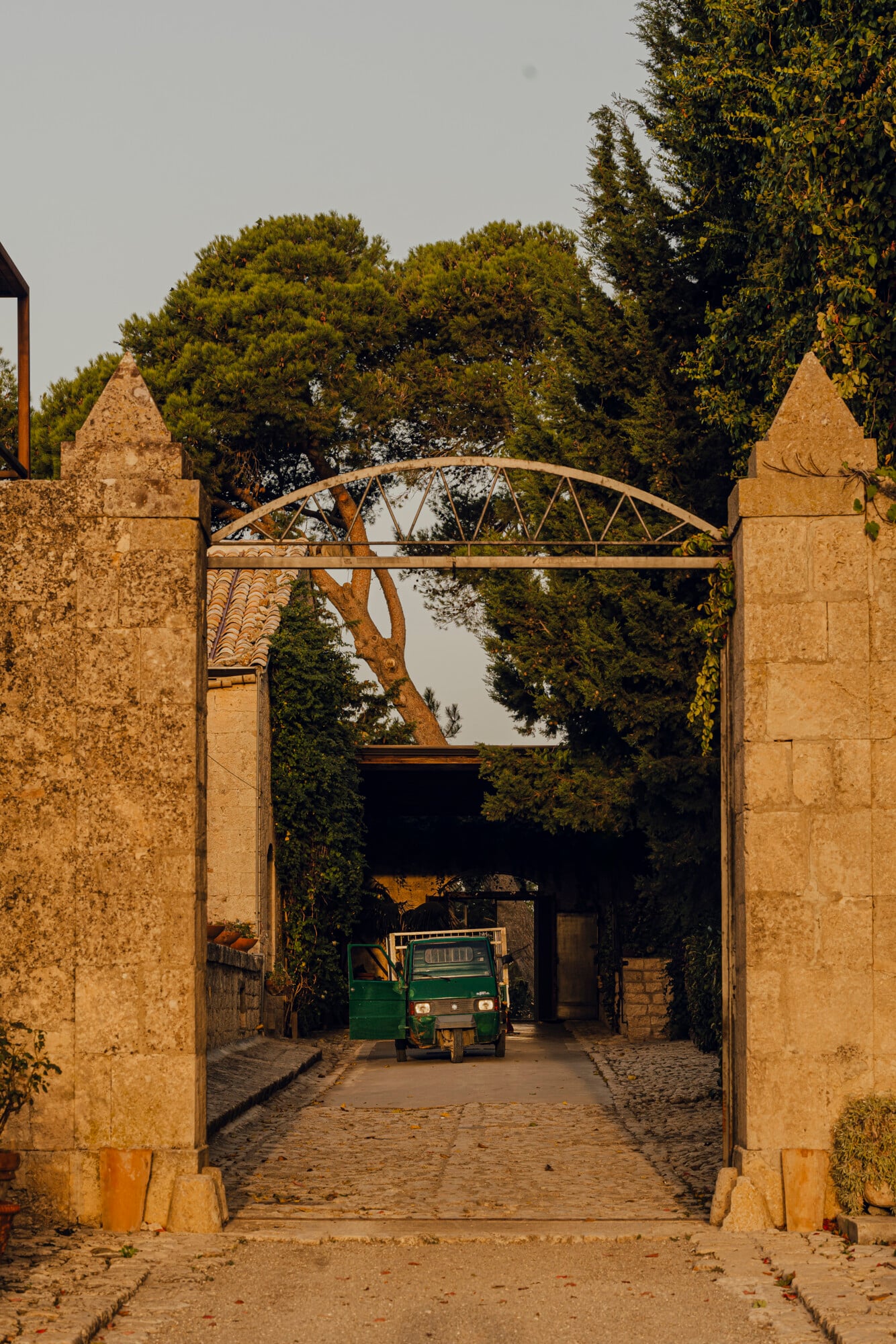 Susafa, Italy — Stone archway with metal gate opening onto cobblestone courtyard, green utility vehicle parked beyond, mature trees and ivy-covered walls framing entrance.