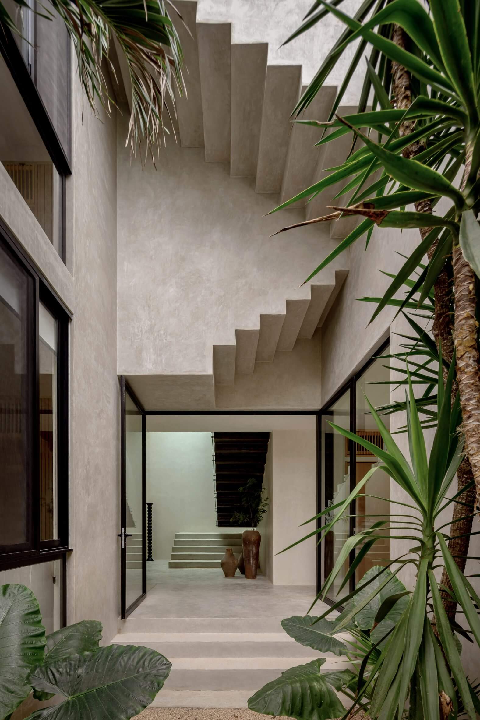 Villa Cava, Mexico — Minimalist courtyard with stepped concrete walls, tropical plants, and floating staircase leading to bright interior spaces.