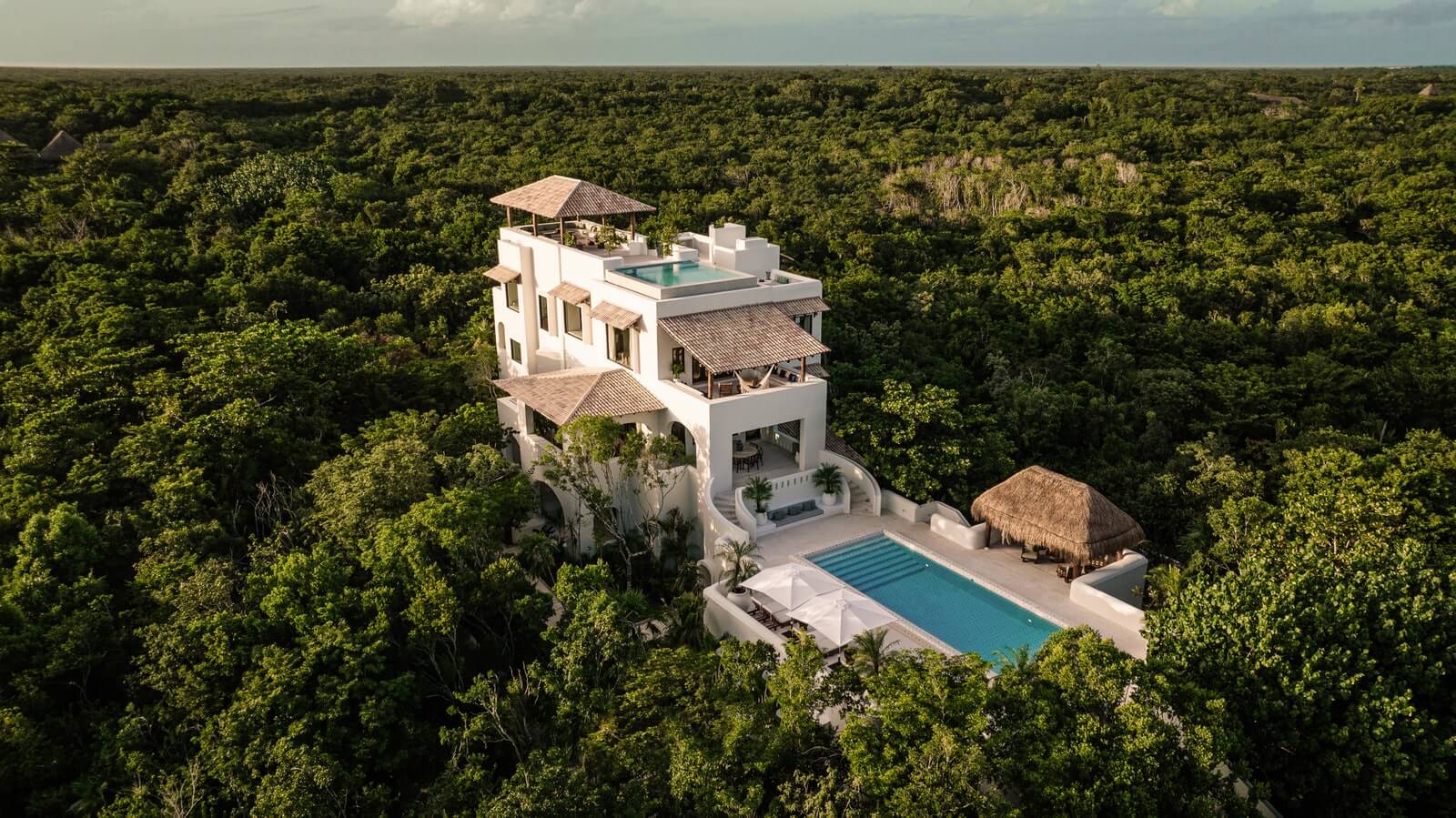 Hotel Esencia, Mexico — Modernist beachfront villa with white stucco walls and tiled roofs rising above dense jungle canopy, infinity pool and palapa structures below.