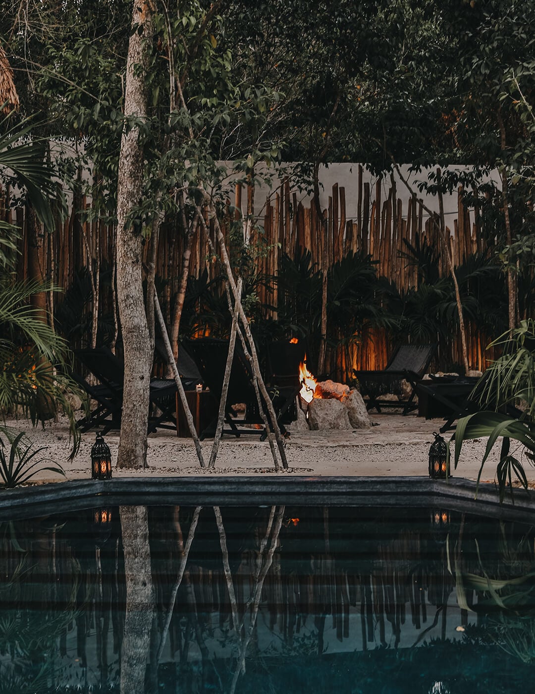Bardo, Mexico — Candlelit courtyard with reflective pool, palm trees, and thatch-roof structures at dusk.