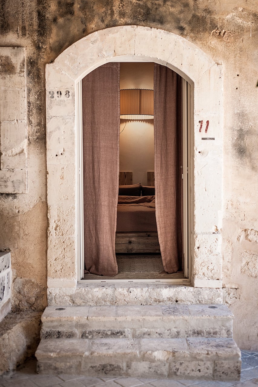 Dimora delle Balze, Europe — Arched limestone doorway with brown linen curtains opening to minimalist bedroom with warm ambient light and wooden furnishings.