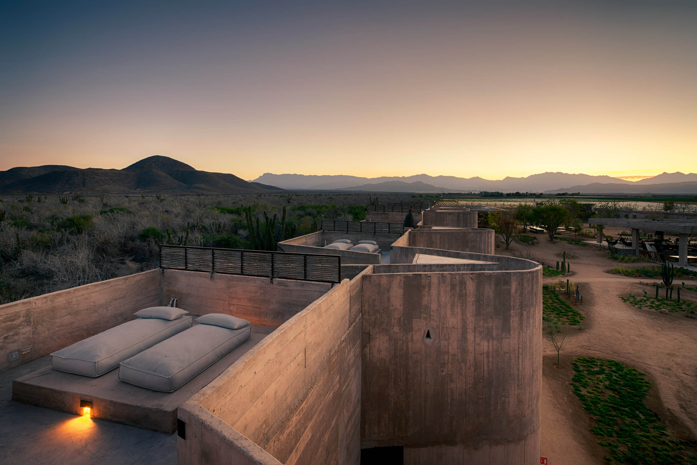 Paradero Todos Santos, Mexico — Desert-view bedroom terrace at sunset with concrete bed platform, wooden privacy screens, and Sierra de la Laguna mountains beyond.