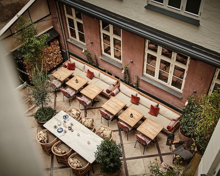 Hotel Sanders, Europe — Courtyard with wooden dining tables, red cushioned chairs, potted plants, and cream stone paving overlooking terracotta facade and tall windows.
