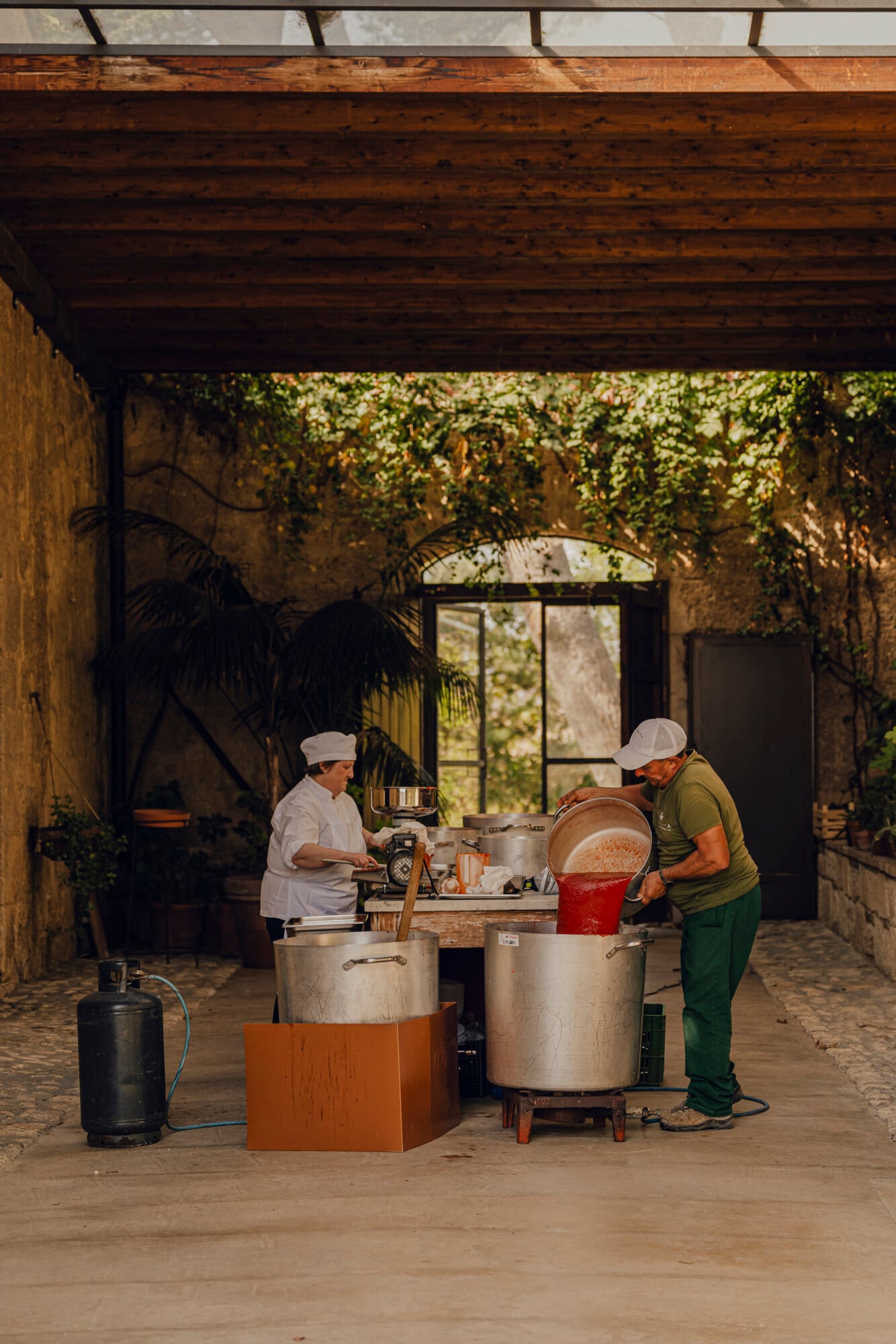Tomato Sauce Class, Italy — Two chefs preparing fresh tomato sauce in a rustic courtyard kitchen with ivy-draped stone walls and dappled natural light.