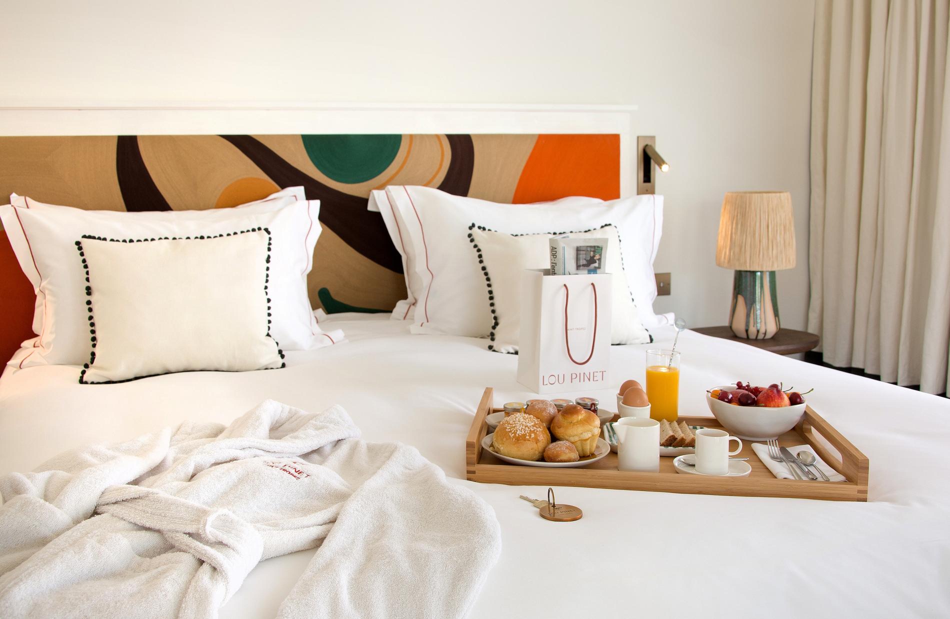 Hôtel Lou Pinet, Europe — Luxury bedroom with geometric headboard, breakfast tray, fresh pastries, orange juice, and geometric patterned pillows in warm earth tones.