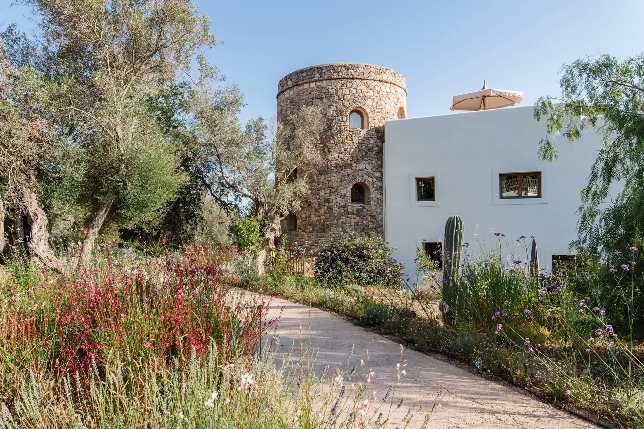 Finca La Inika, Balearic Islands, Spain — Stone watchtower adjoining whitewashed modern villa with Mediterranean garden of flowering plants and olive trees.